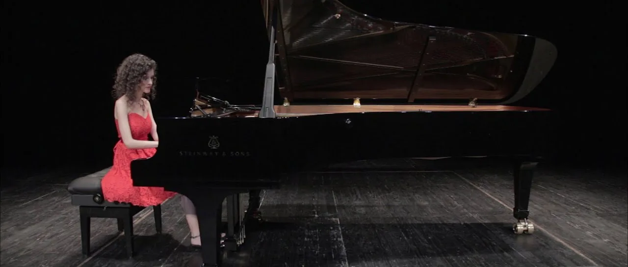 Alicia Sevilla, de rojo, al piano de cola sobre un escenario a oscuras